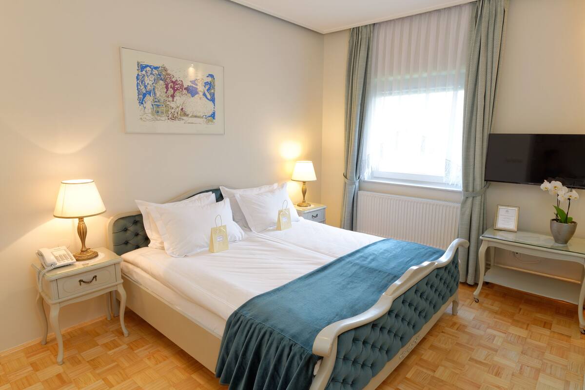A neatly arranged hotel room with a bed, bedside lamps, a bedside table with a lamp, a small desk with a TV, and a window with curtains.