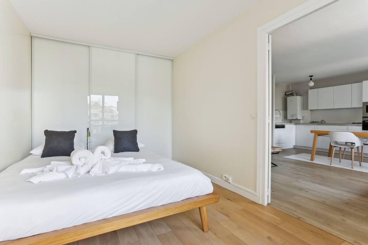 A modern bedroom with a wooden bed, white bedding, and dark pillows. A bathroom is visible in the background.