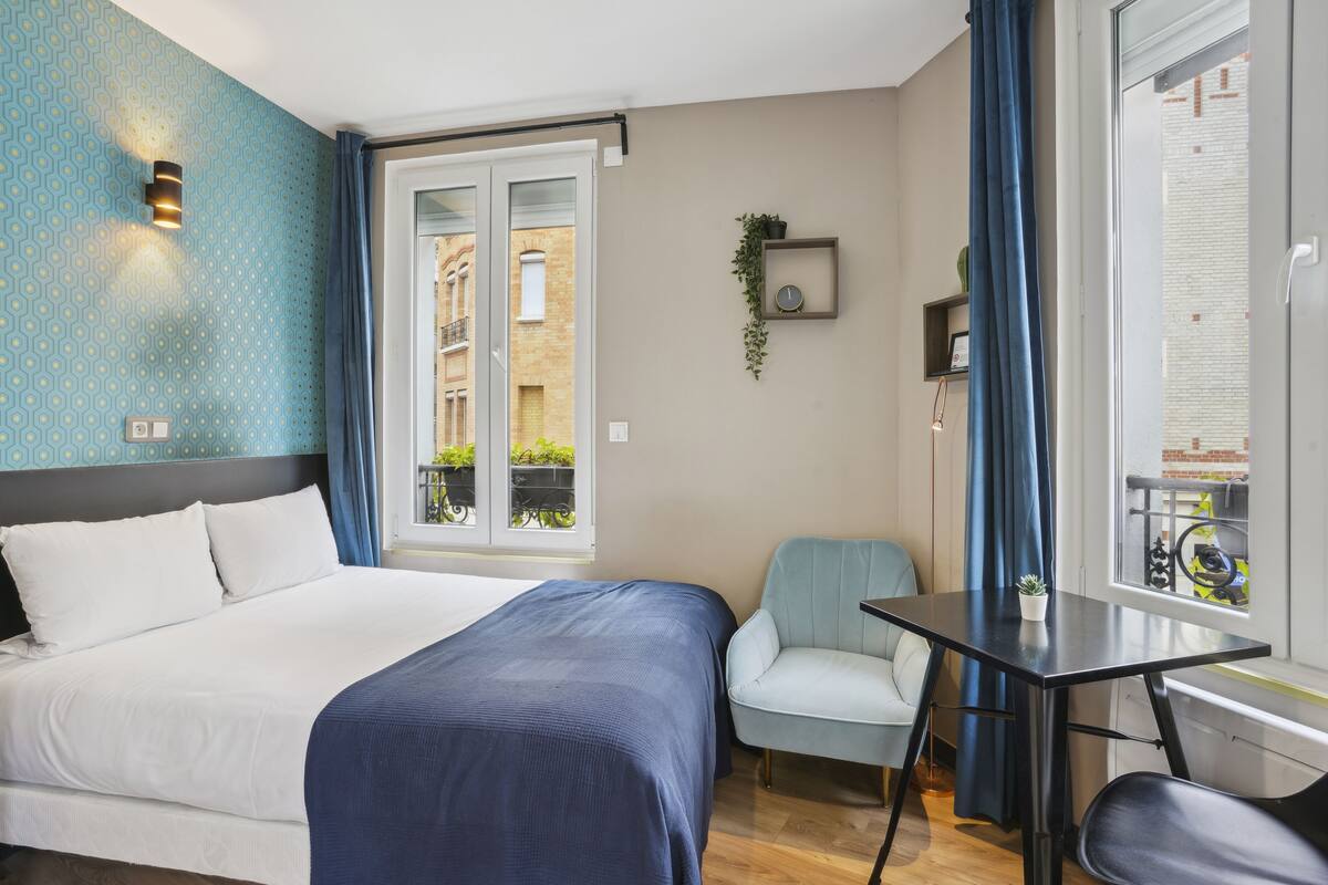 A hotel room with a bed, a small table, a chair, a window with curtains, and a wall-mounted clock.
