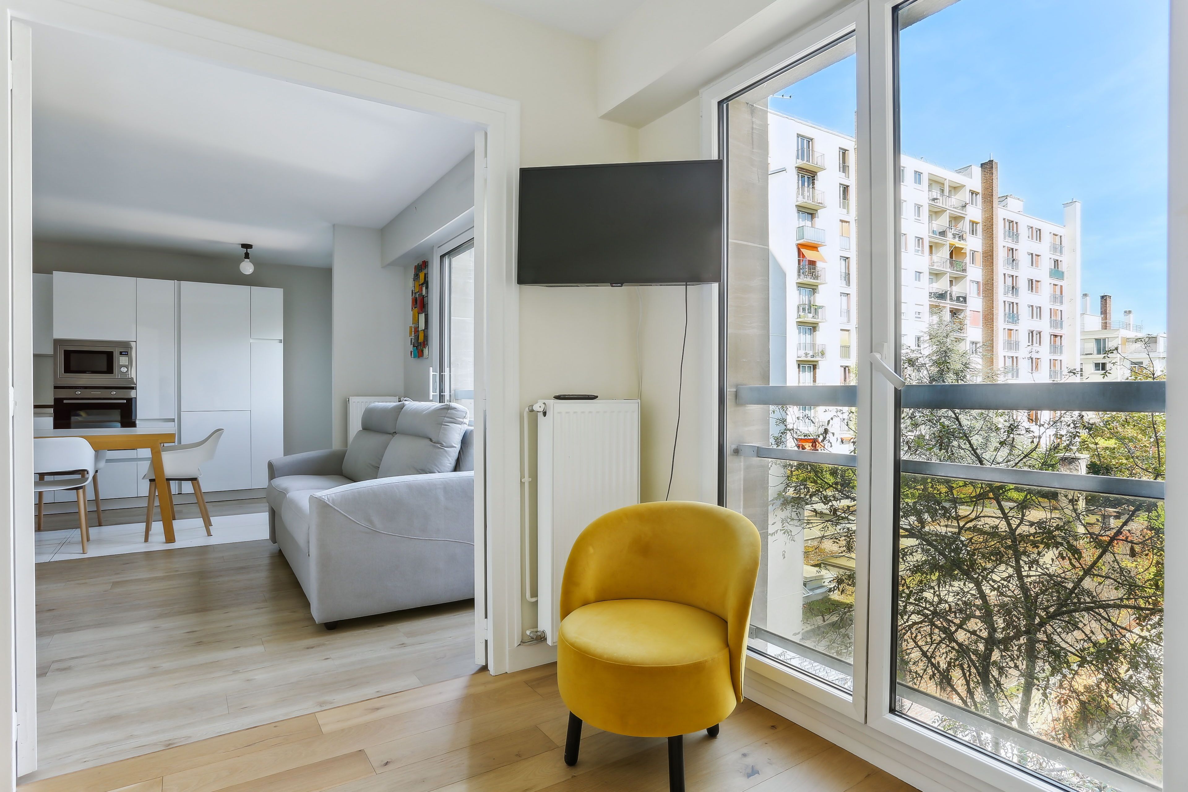 A modern apartment with a living area, kitchen, and dining space. It features a large window with a view of a residential building.