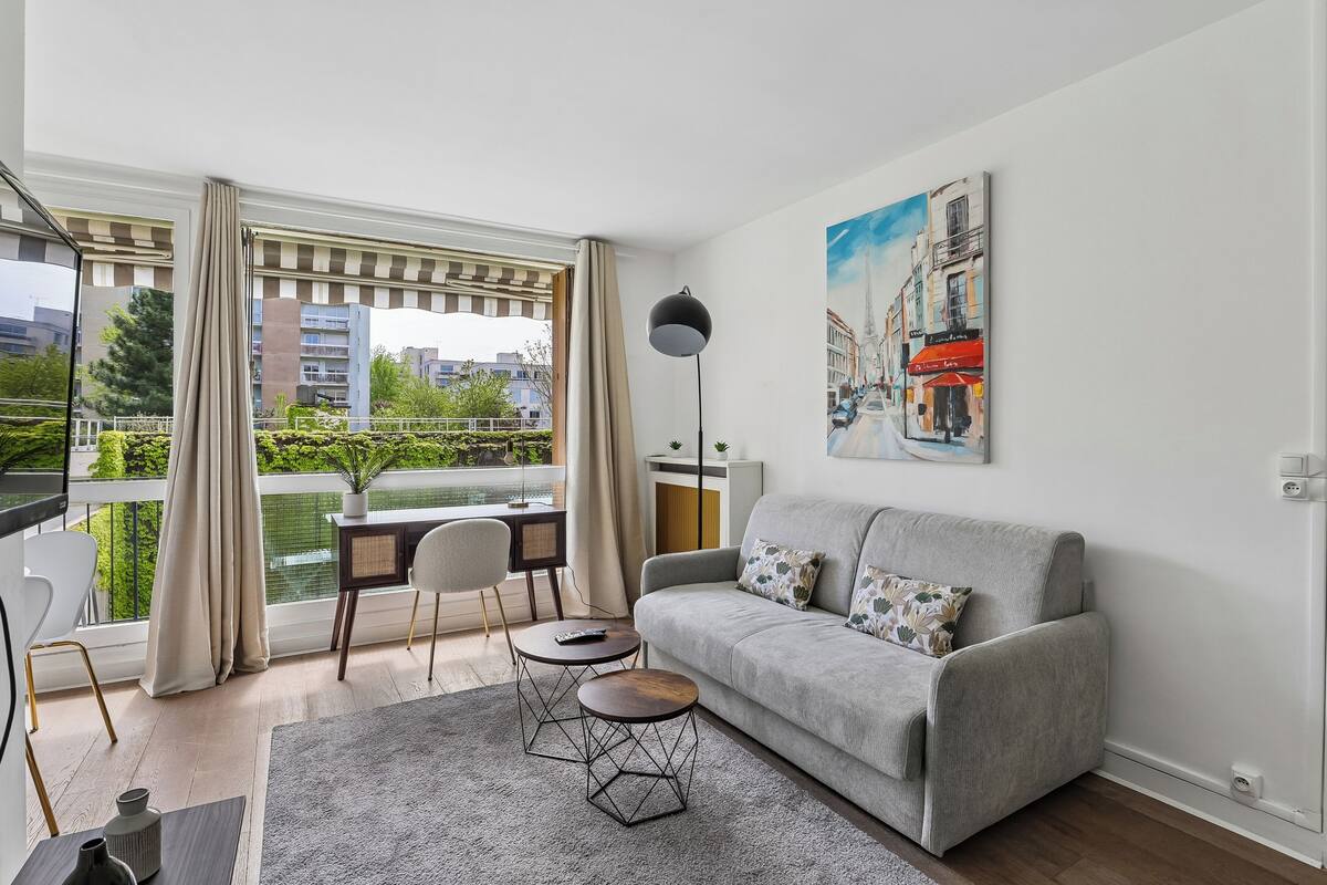 A modern living room with a sofa, two small tables, a large window with curtains, a painting, and a view of buildings.