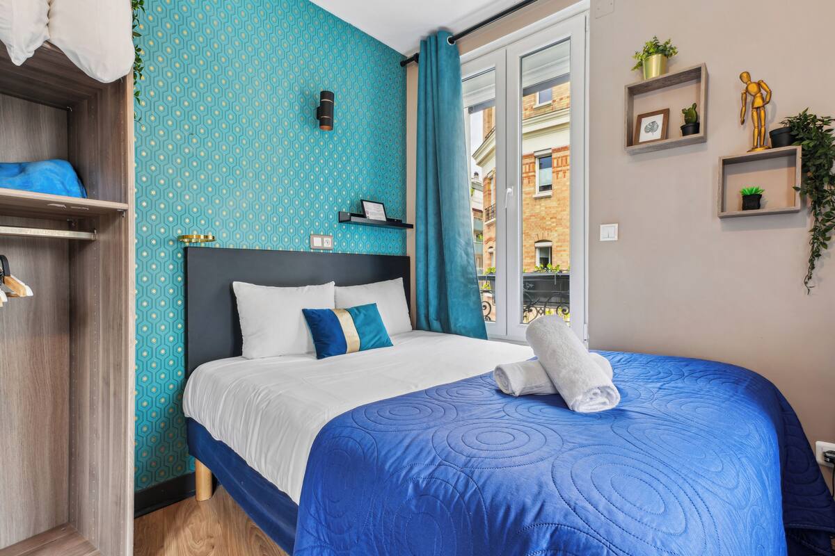 A neatly made bed with a blue quilt, a wooden wardrobe, and a view of the street through a window with curtains.