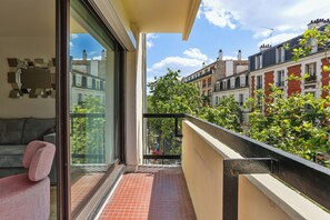 A balcony with a red-tiled floor, a pink chair, and a glass door leading to a living area. - Résidence Aurmat Appart Hôtel Paris Boulogne (Boulogne-Billancourt)