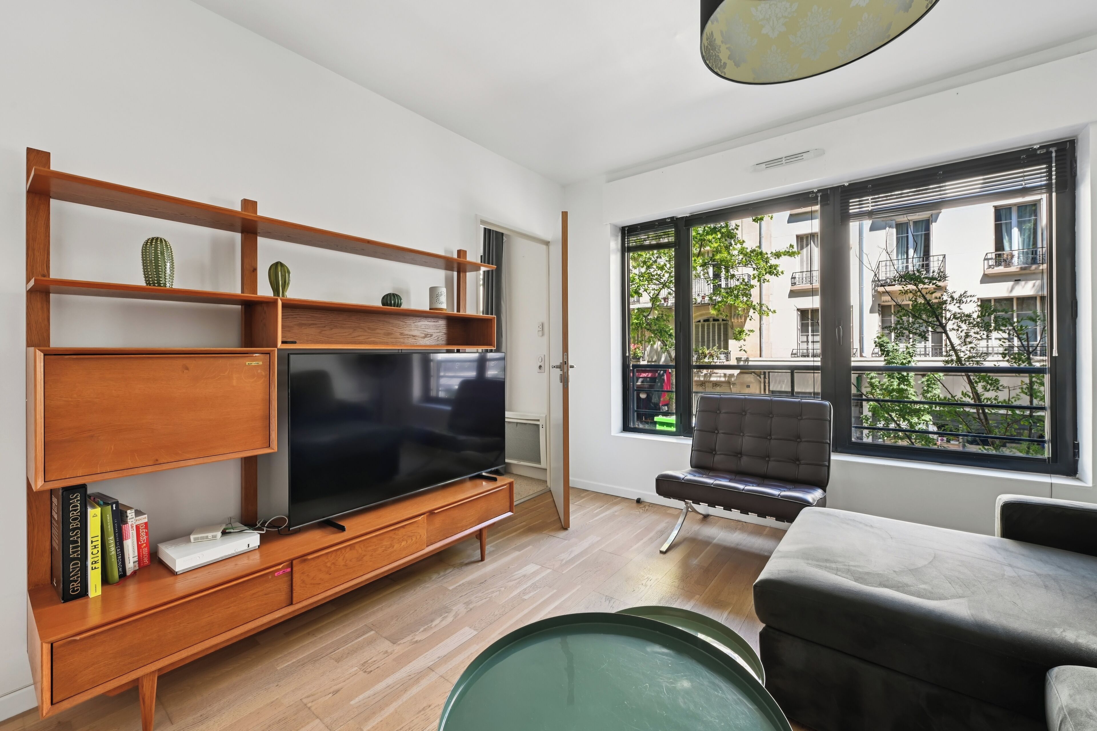 A modern living room with a flat-screen TV, wooden shelves, a bench, and a large window with a view of buildings.