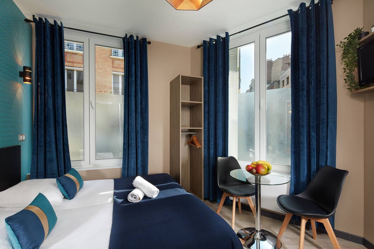 A hotel room with a bed, a desk with a chair, a small table with fruit, and a wardrobe.