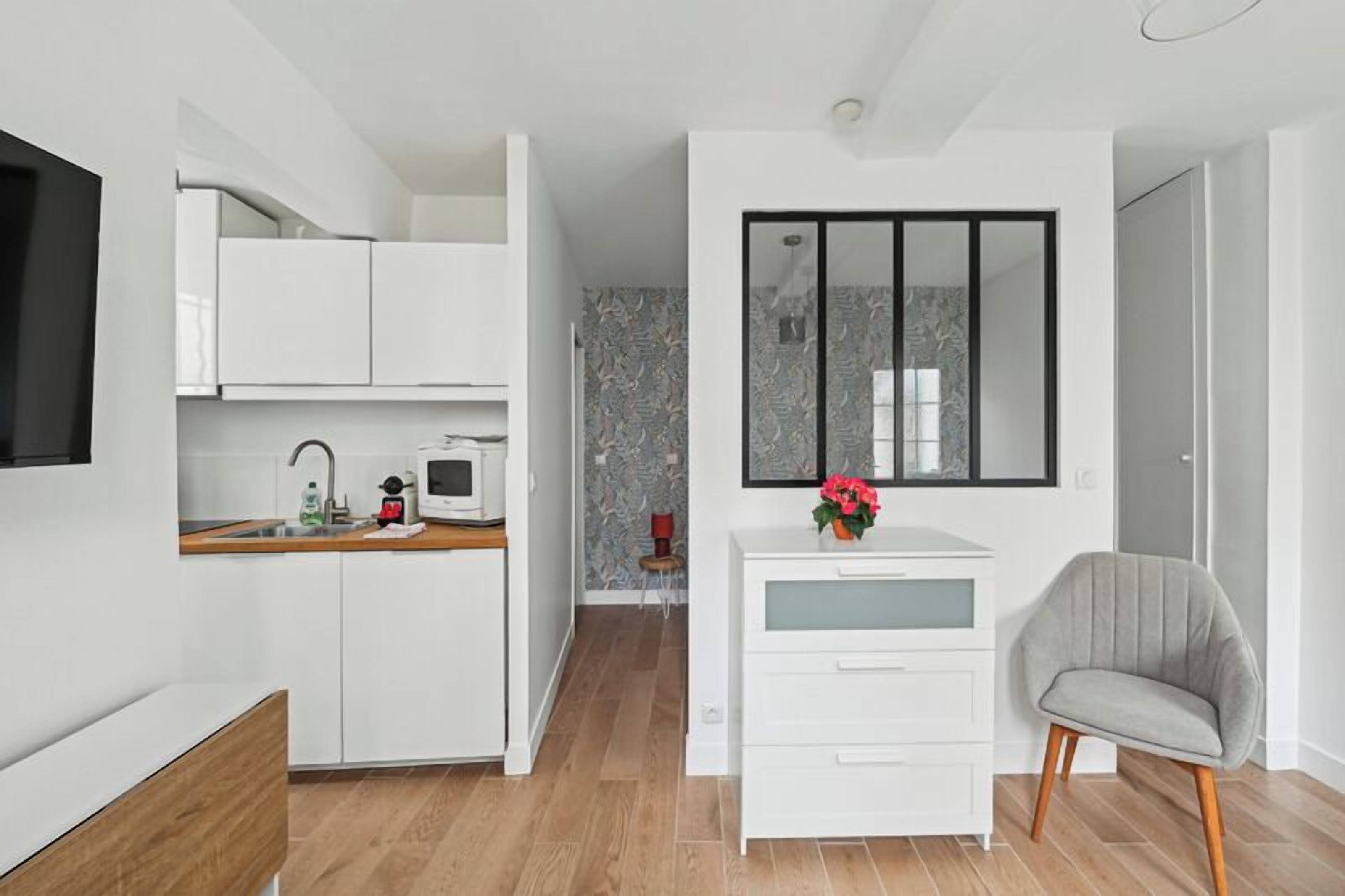 Una cocina moderna con armarios blancos, un microondas y un lavamanos. Hay una pequeña mesa con una silla y un jarrón con flores.