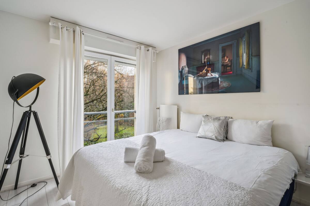 A neatly made bed with white linens and a rolled towel, a tripod lamp, and a painting of a room scene on the wall.