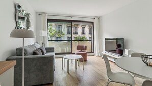 A modern living room with a sofa, a round table, a TV, and a large window with a view of buildings. - Résidence Aurmat Appart Hôtel Paris Boulogne (Boulogne-Billancourt)