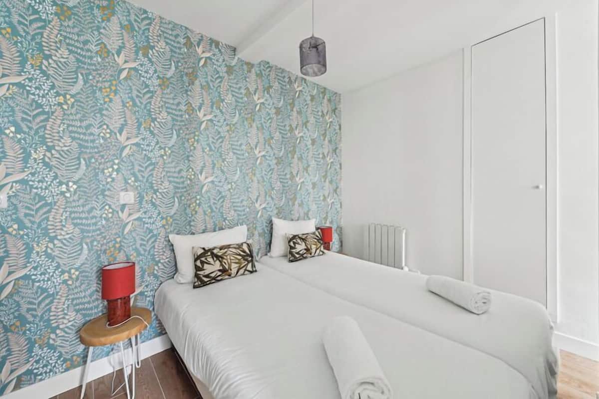 A modern bedroom with a patterned wallpaper, a bed with white linens, a red lamp on a wooden side table, and a radiator.