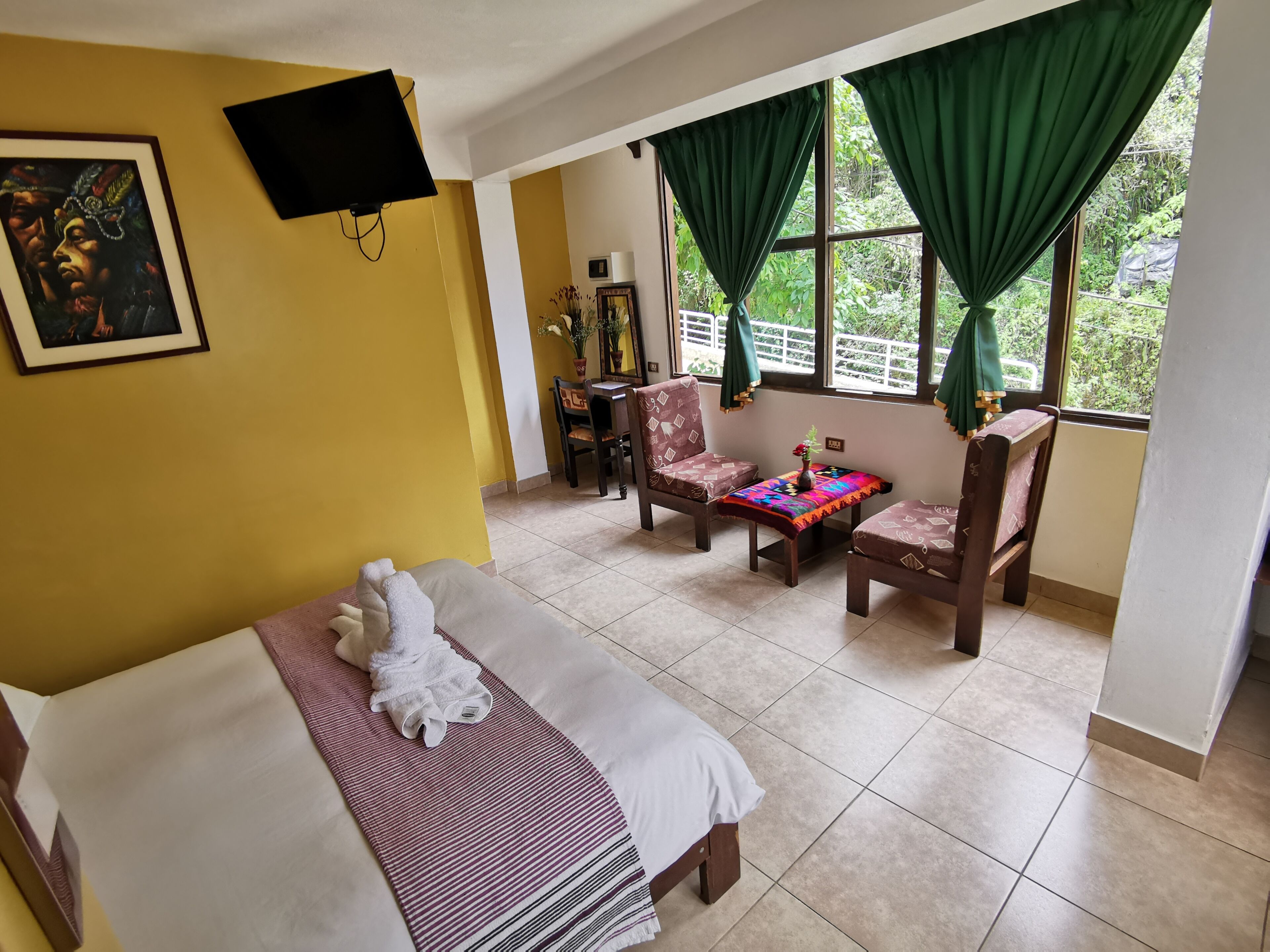 Una habitación de hotel con una cama, dos sillas, una mesita, un televisor y un ventanal con cortinas.