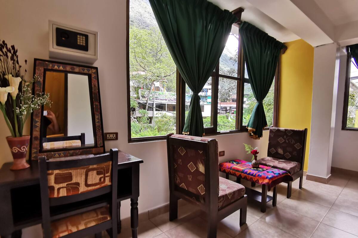 A room with a wooden desk, a chair, a bed with a colorful blanket, and a window with green curtains.