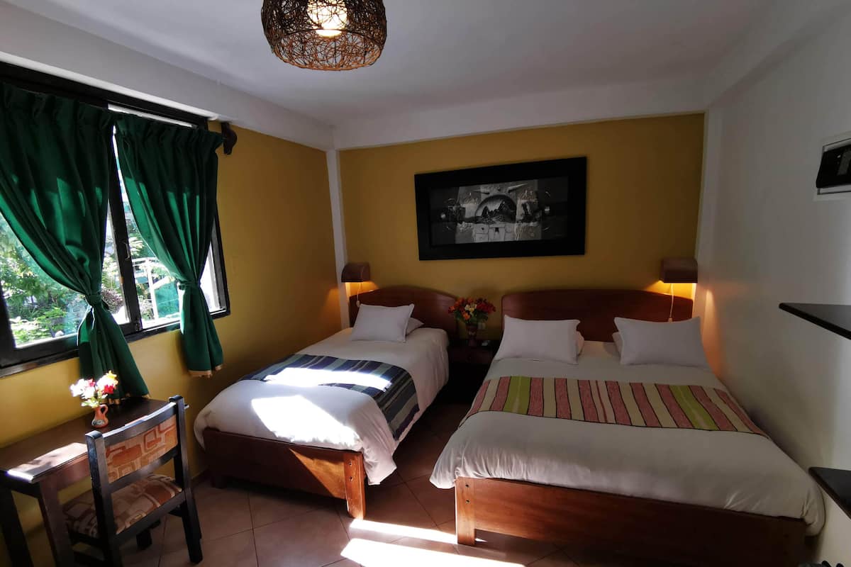 A hotel room with two beds, a chair, a table, a wall-mounted picture, and a window with curtains.