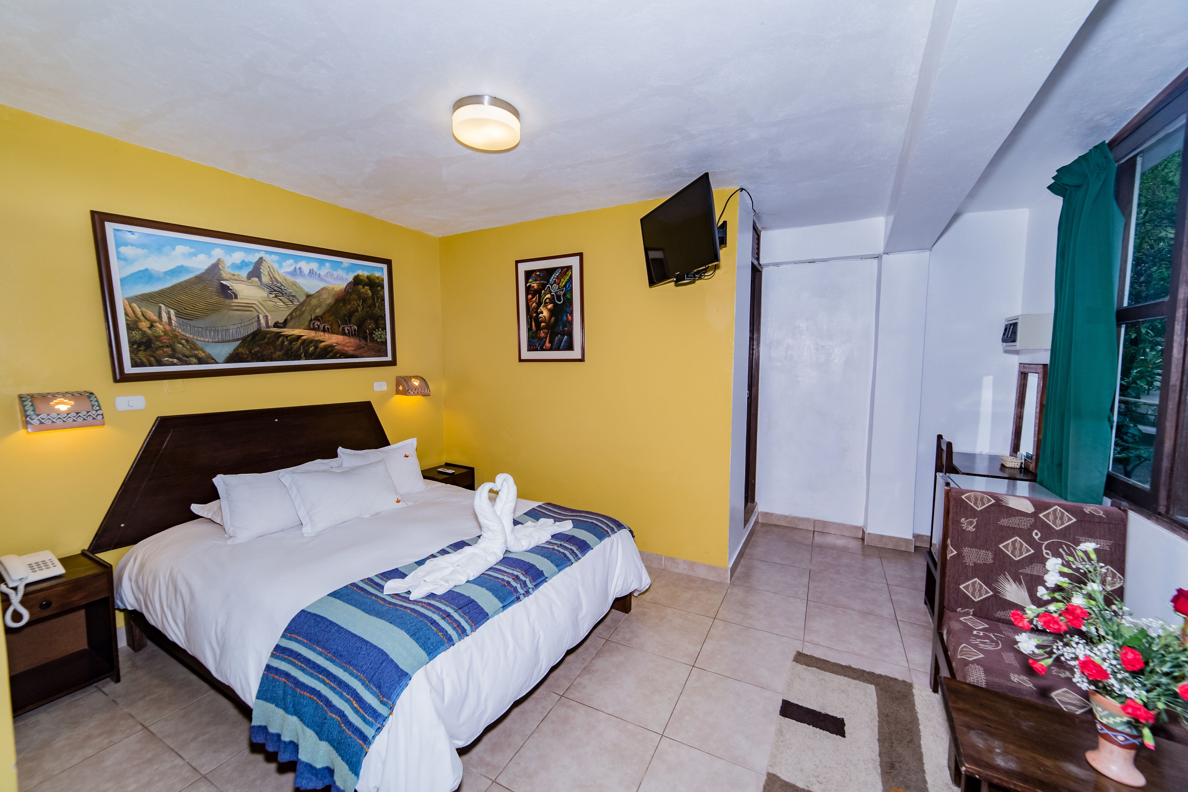 A hotel room with a bed, a nightstand, a telephone, a painting on the wall, a television, and a window with curtains.