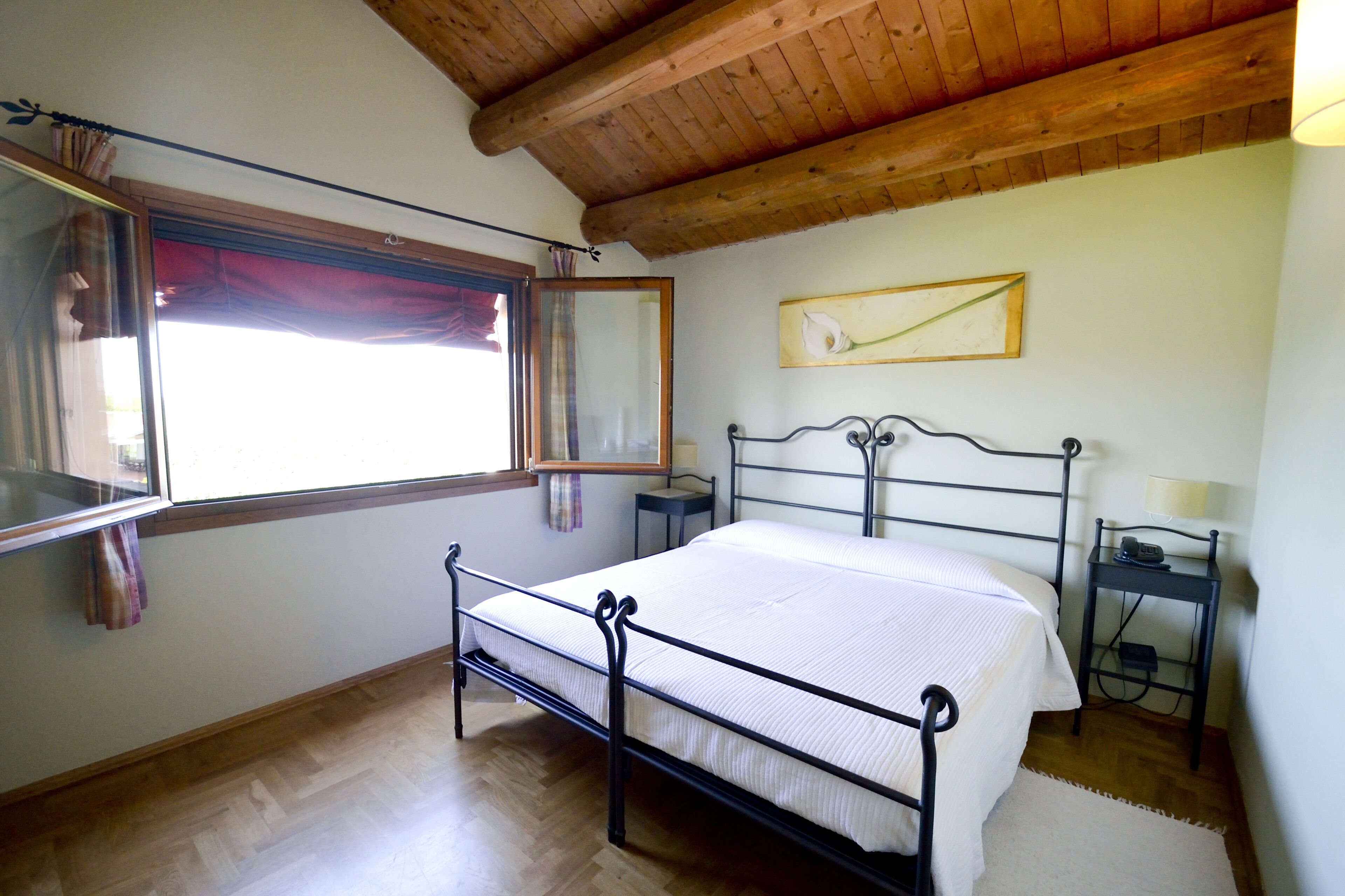 A bedroom with a wooden ceiling, a bed with a metal frame, a window with curtains, a bedside table, and a wall-mounted lamp.