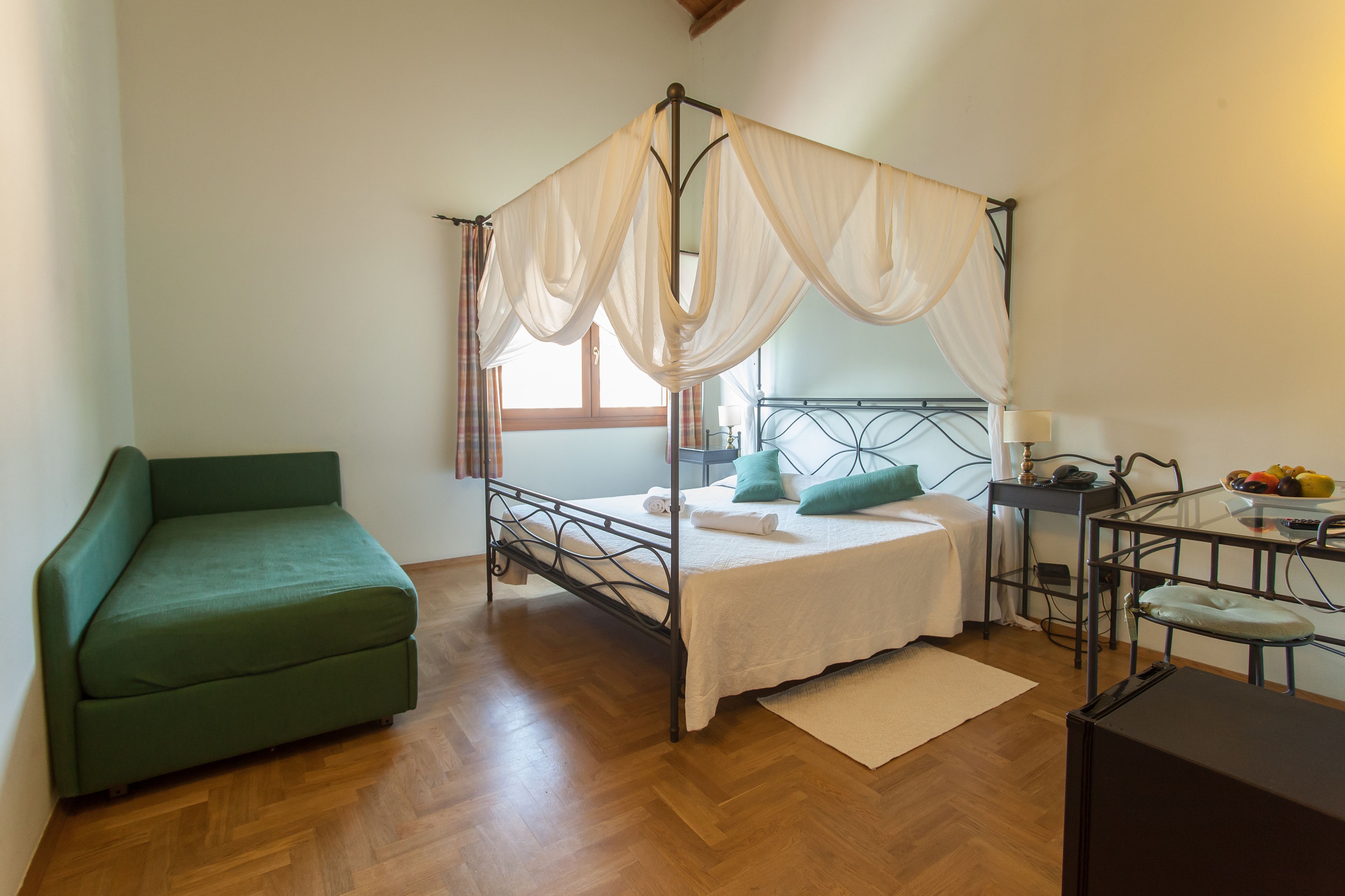 A room with a four-poster bed, a green sofa, a small table with a lamp, and a fruit bowl.