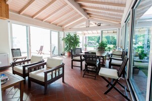 A sunlit patio with wooden furniture and large windows offering a view of the outdoors.