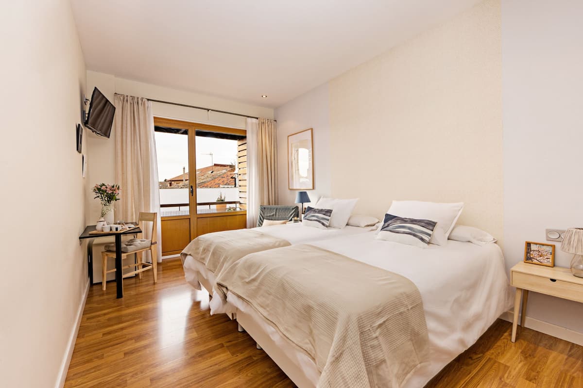 Quarto com duas camas, uma escrivaninha, uma cadeira e janela com vista para edifícios.