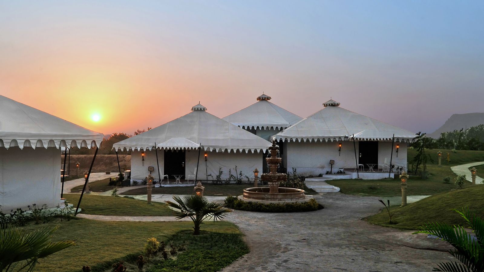 A series of yurts with a central fountain, surrounded by a landscaped garden, during sunset.