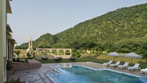 Une piscine entourée de chaises longues et de parasols, elle-même bordée d’un jardin aménagé et avec une colline en arrière-plan.