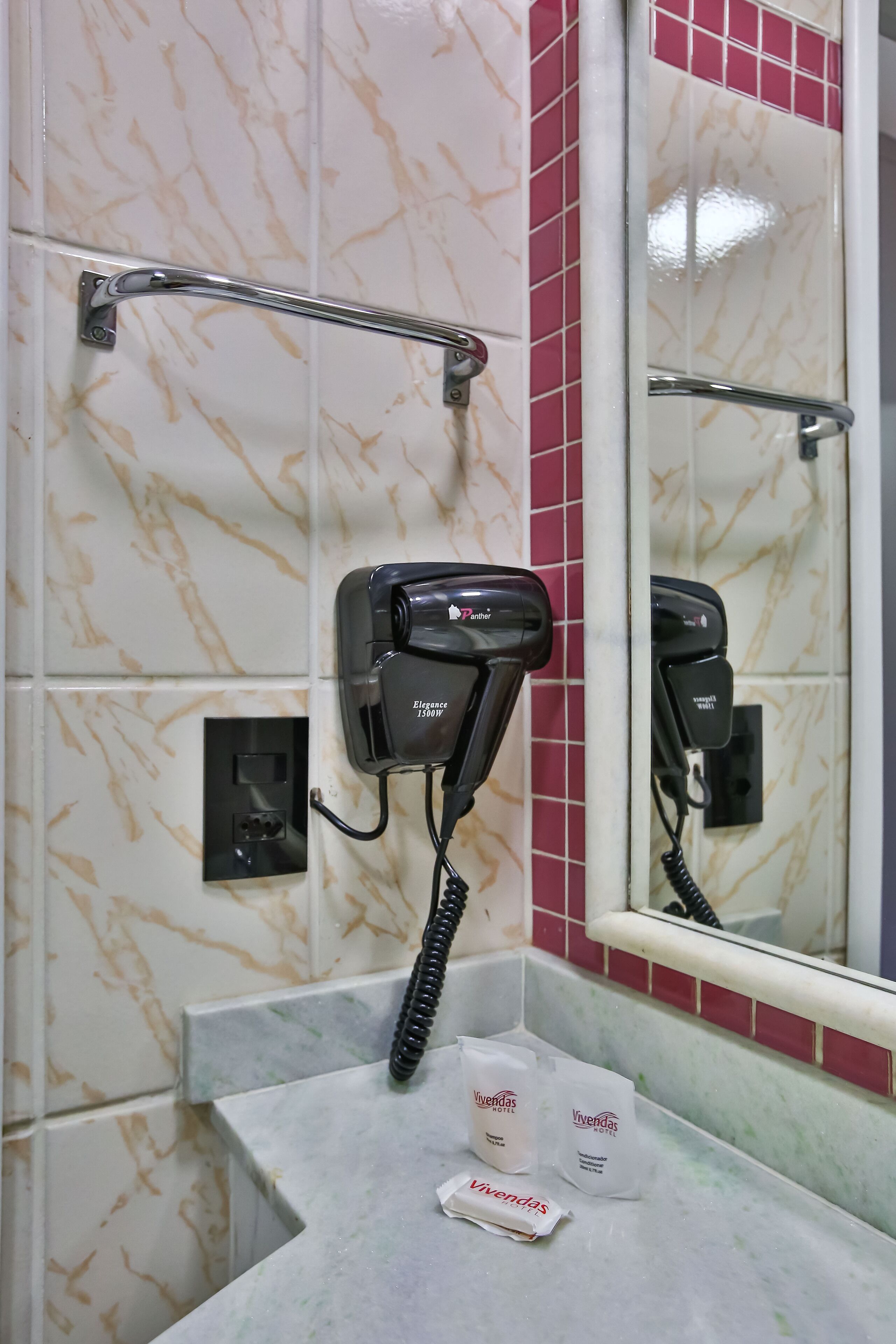 A bathroom with a hairdryer mounted on the wall, a towel rack, and toiletries on the counter.