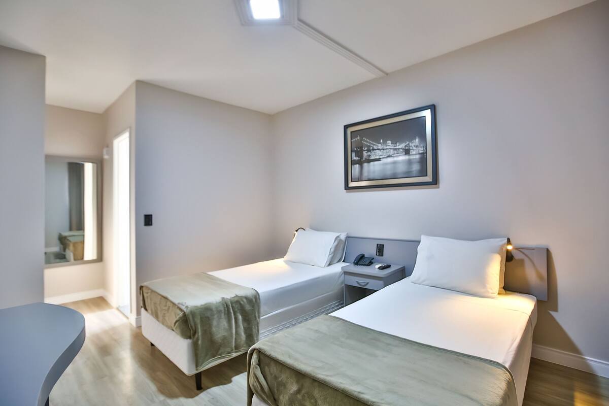 A hotel room with two beds, a desk, a chair, a framed picture on the wall, and a window.