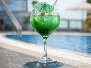 A glass of green drink with a straw and a green umbrella on a poolside.