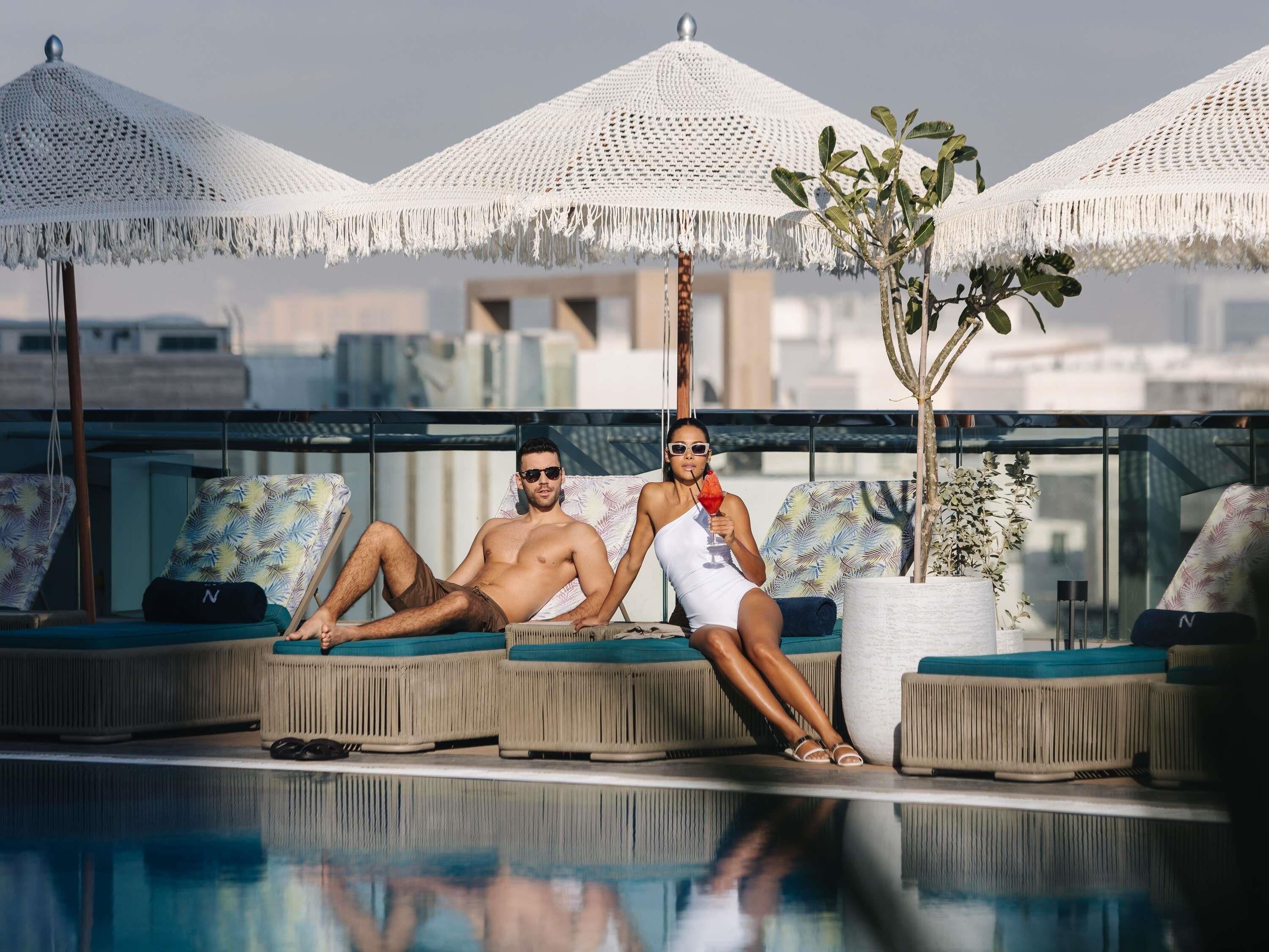 A poolside area with lounge chairs and umbrellas, a clear view of the sky, and a reflection in the water.