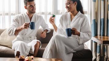 A couple in white bathrobes sitting on a couch with a tray of food and drinks.