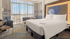 Une chambre d’hôtel avec un grand lit, un bureau, une chaise et une vue sur la ville grâce à de grandes fenêtres.
