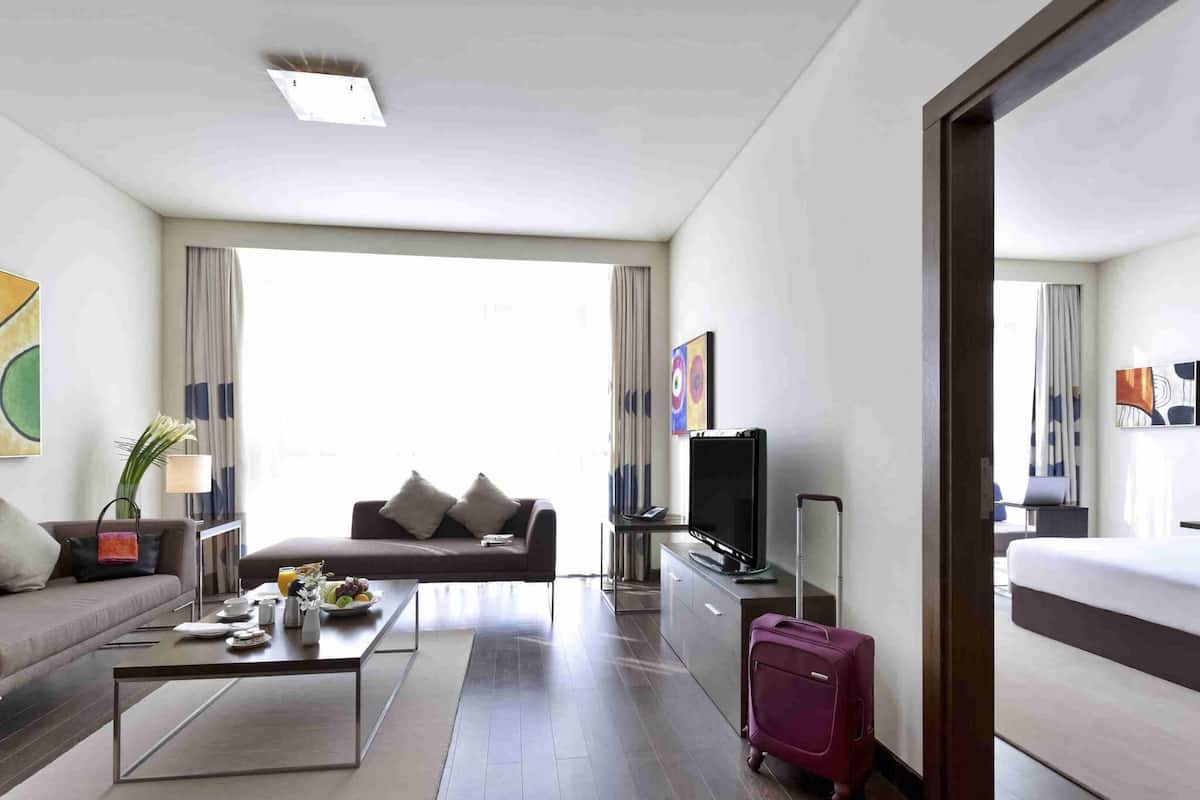 A hotel room with a sofa, a coffee table, a TV, a suitcase, and a bed.