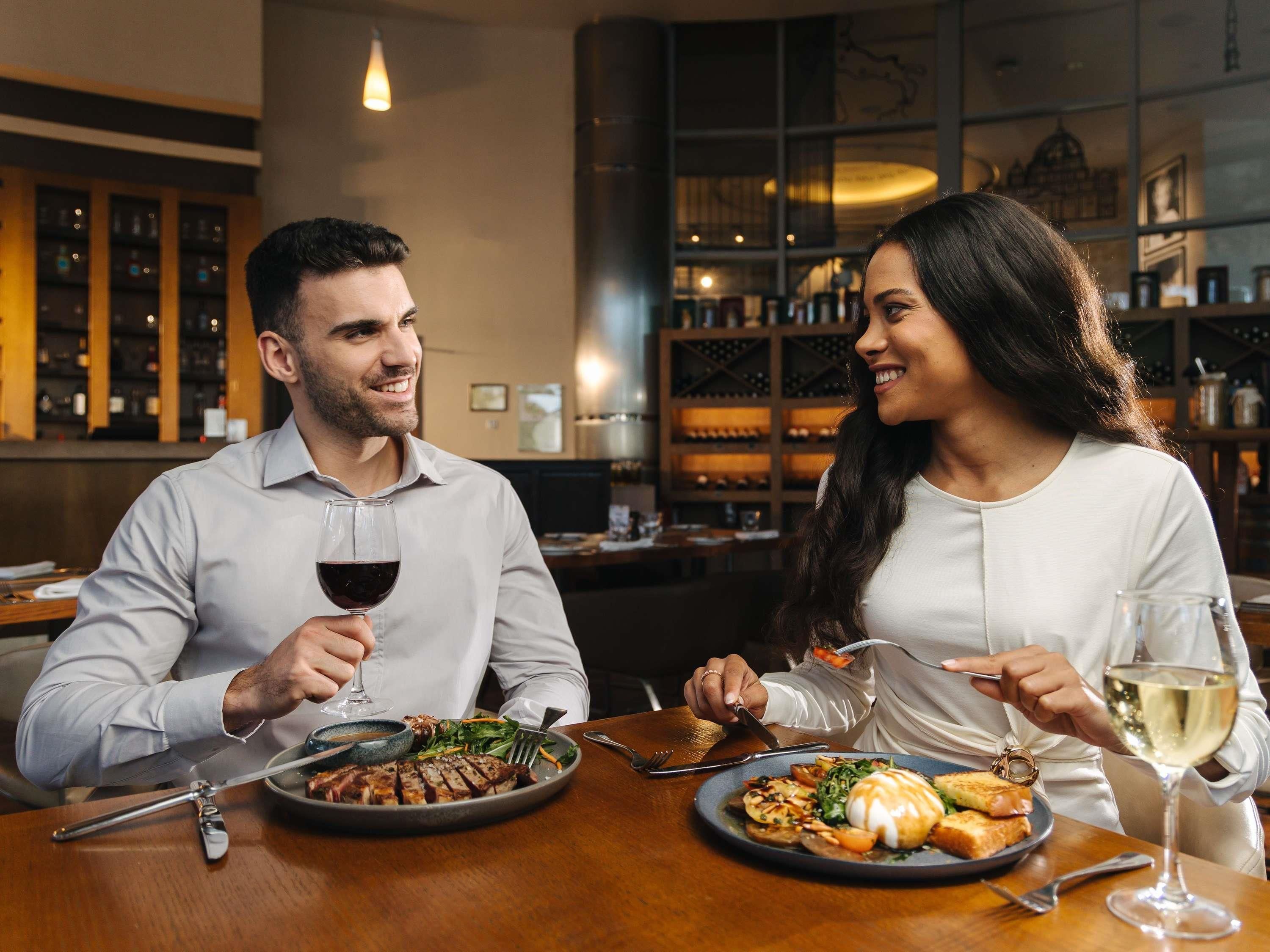 Un couple est assis à une table dans un restaurant, avec un verre de vin et des assiettes remplies de nourriture.