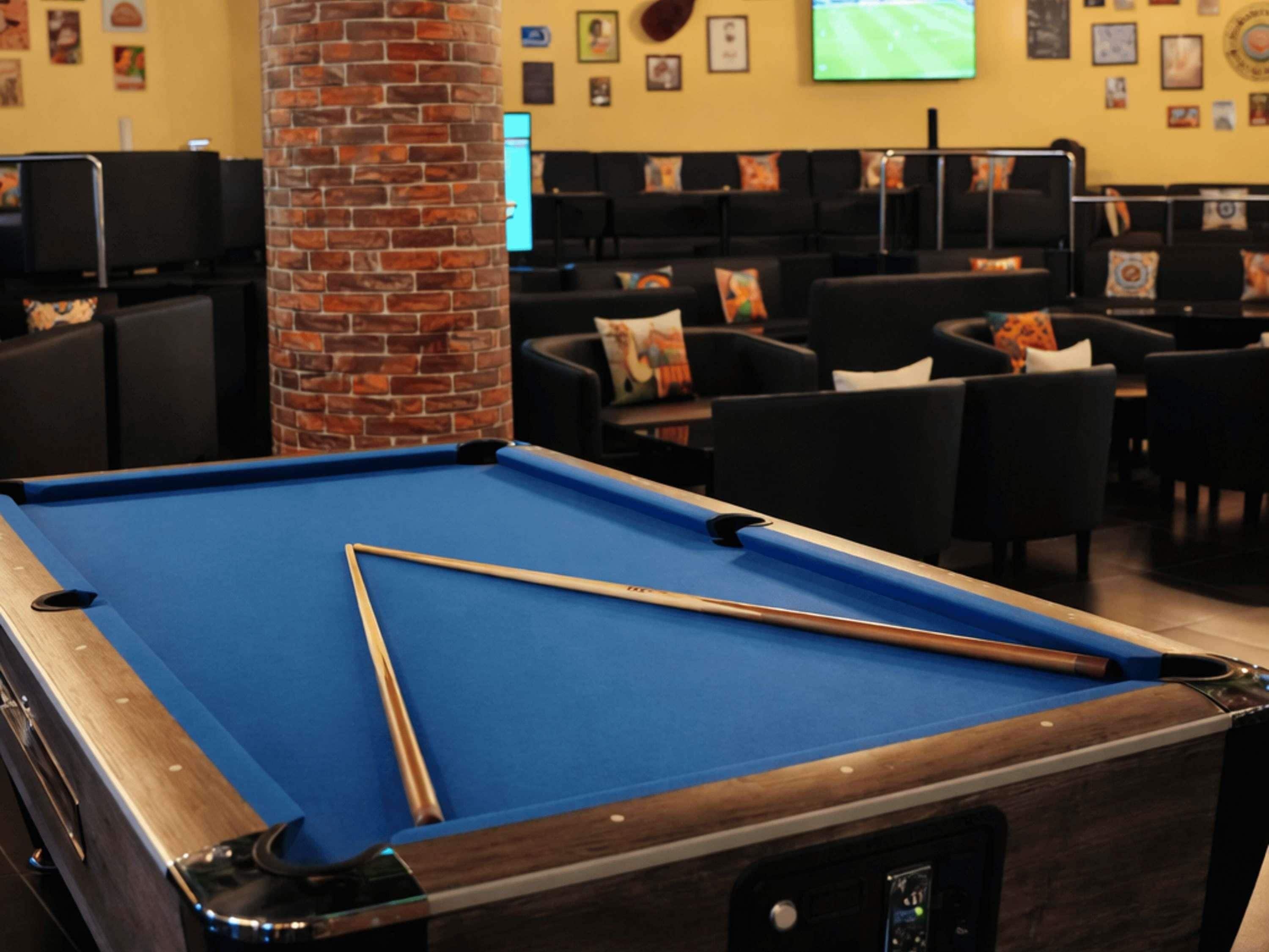 A pool table with a wooden frame and blue felt, surrounded by black chairs and a brick pillar.