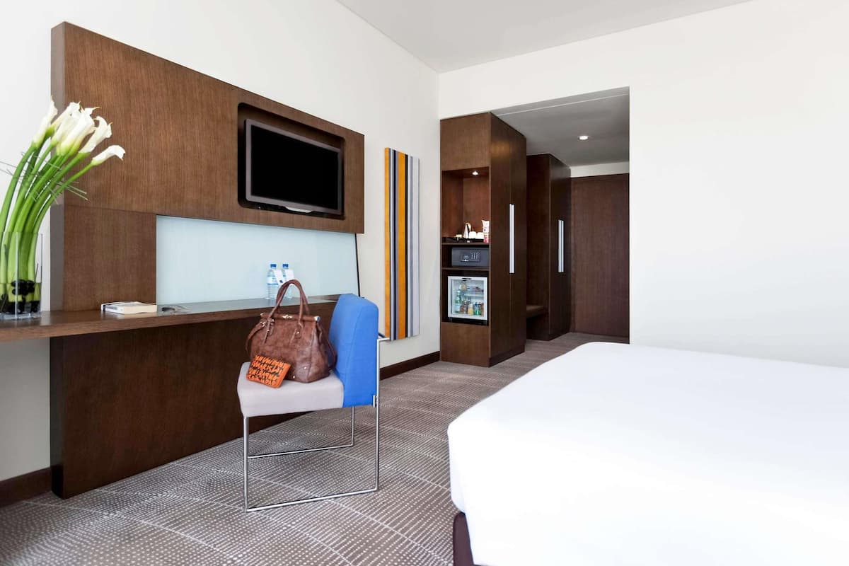 A hotel room with a bed, a desk with a TV, a chair, a minibar, and a door leading to another room.