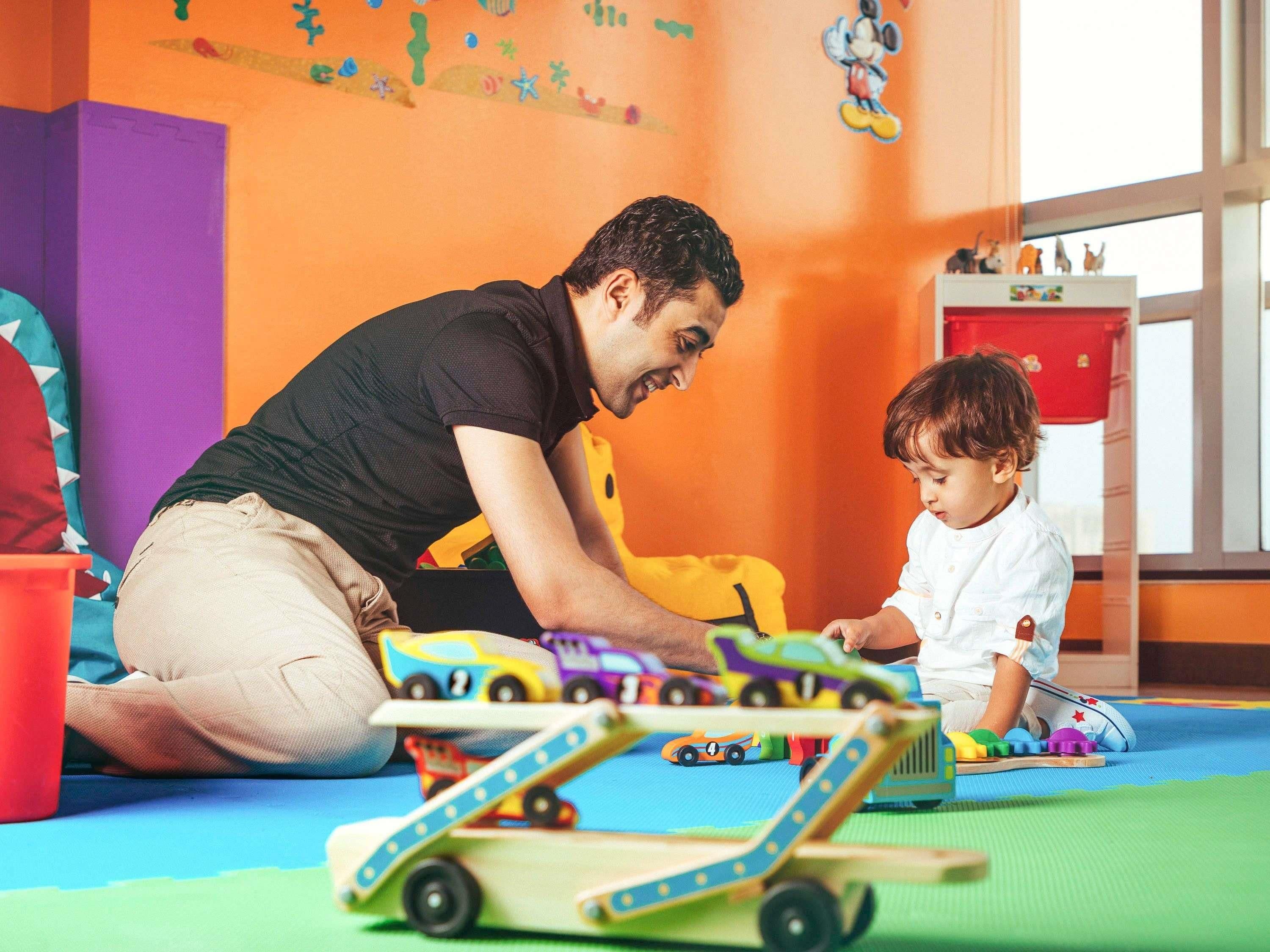 A man and a child playing with toy cars on a colorful play mat in a room with orange walls and a window.