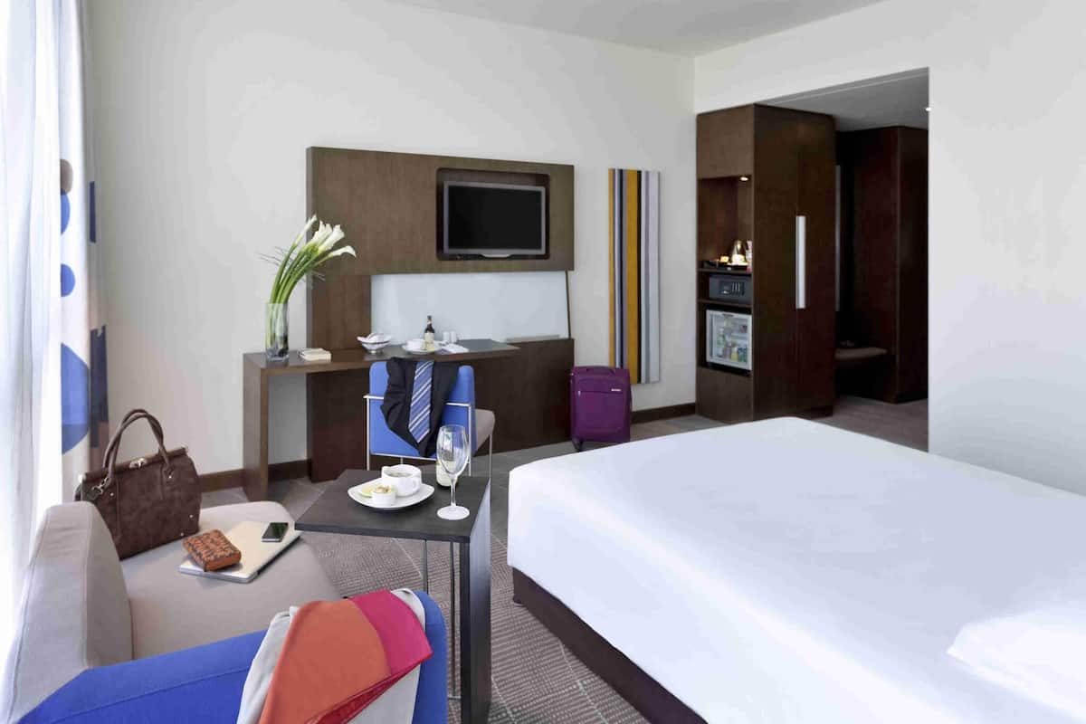 A hotel room with a bed, a desk with a chair, a TV, a minibar, and a bathroom.