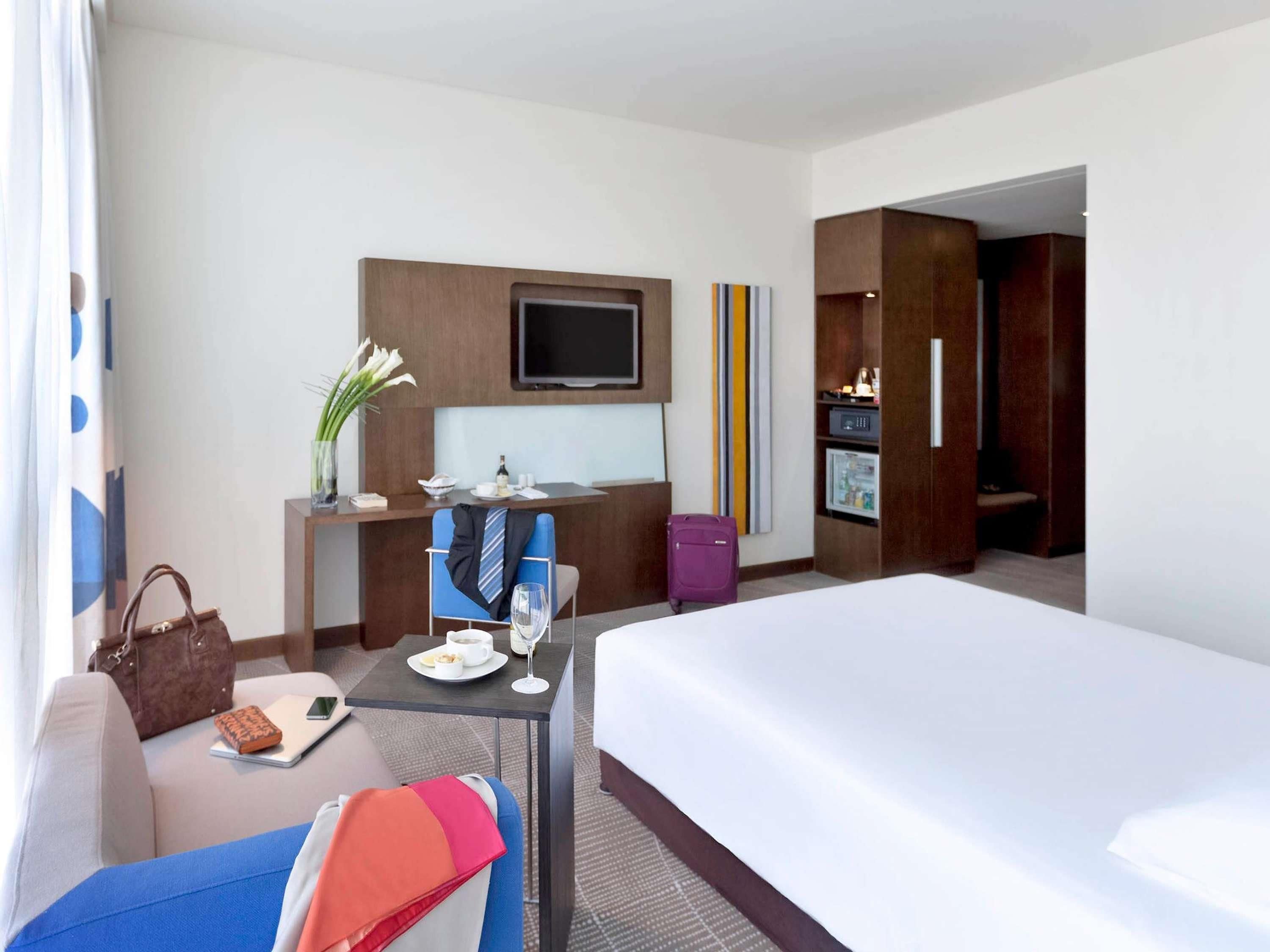 A hotel room with a bed, a desk with a chair, a television, a minibar, and a suitcase.