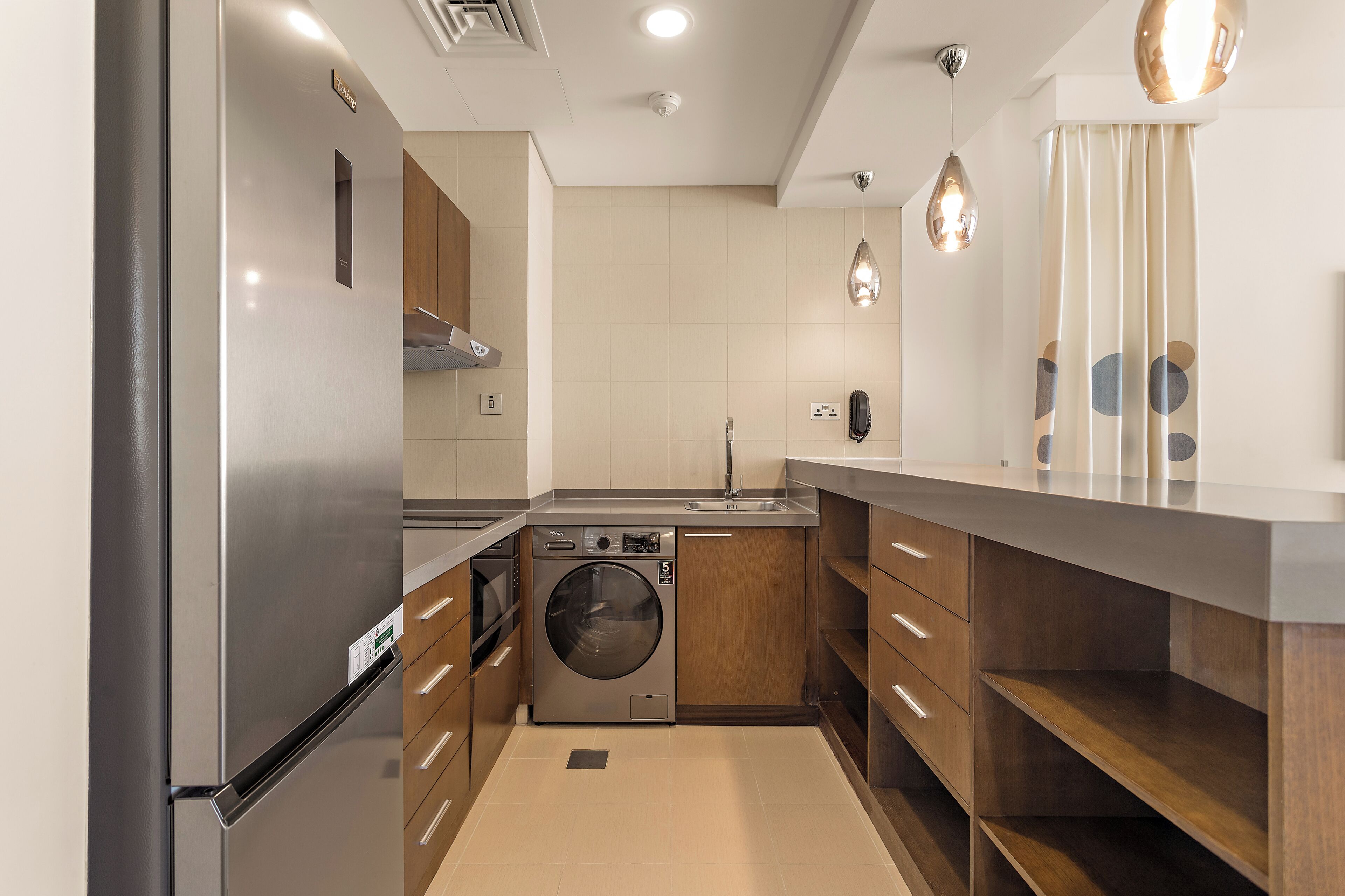 A compact kitchen with a stainless steel refrigerator, a built-in washing machine, wooden cabinets, and a modern light fixture.