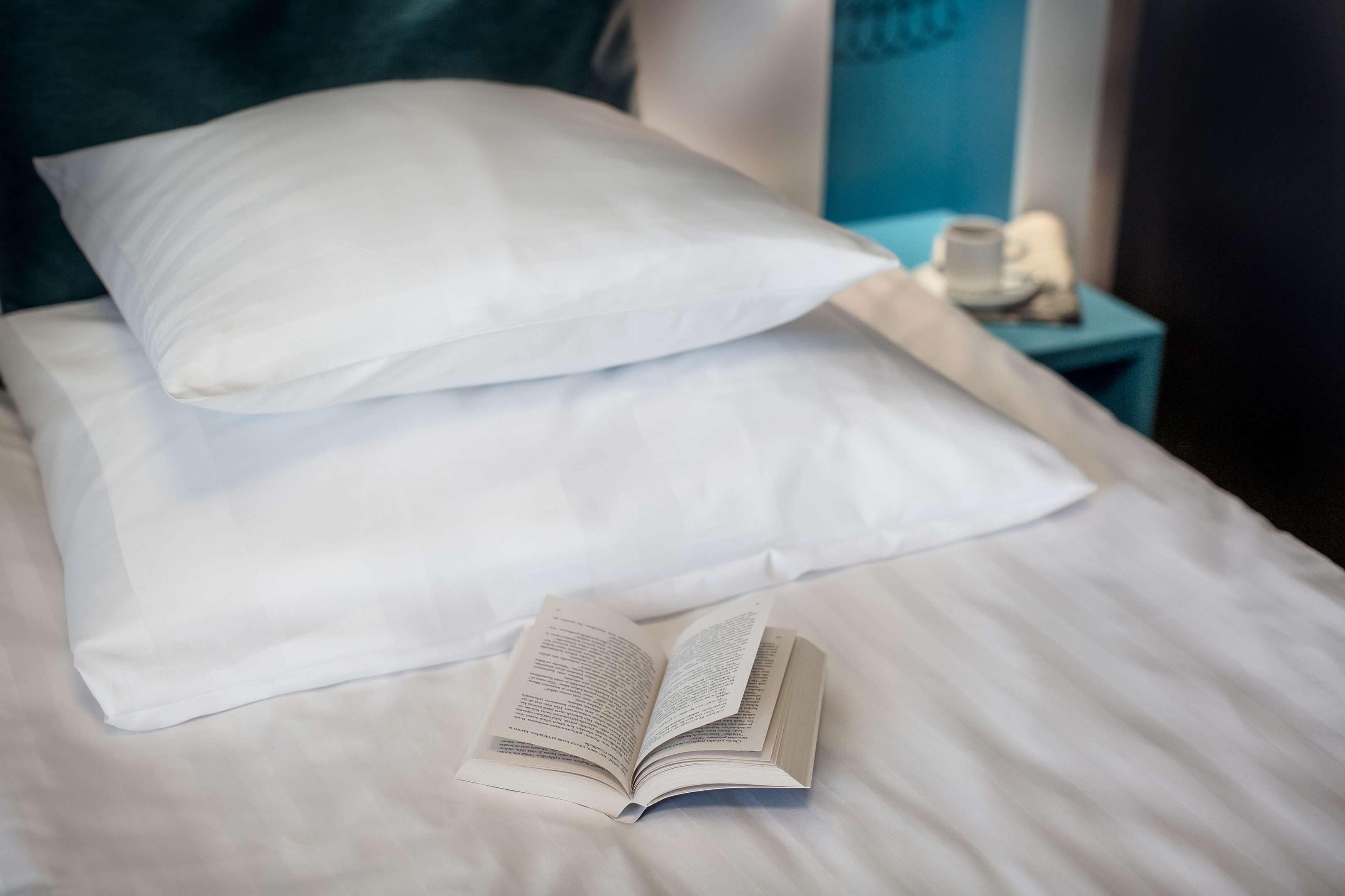 Una cama con sábanas y almohadas blancas, un libro abierto sobre la cama y una mesita de noche con una lámpara y una taza al fondo.