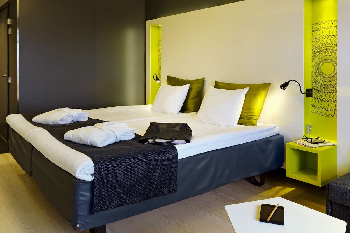 A modern hotel room with a neatly made bed, a bedside table with a lamp, and a small green nightstand.