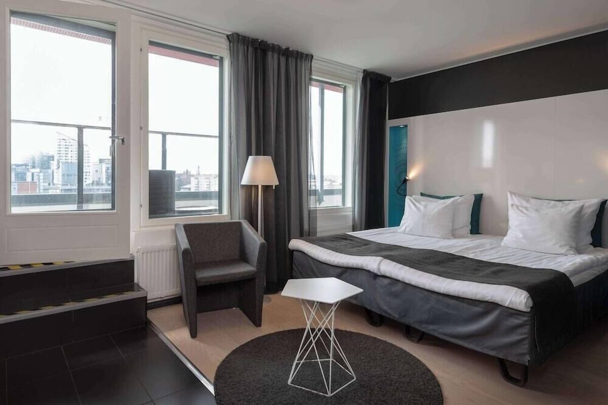 A modern hotel room with a large bed, a chair, a small table, and a view of the city through large windows.
