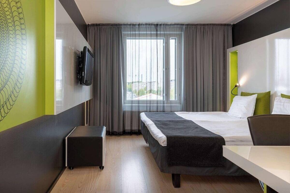A hotel room with a bed, a desk, a chair, a TV, and a window with curtains.
