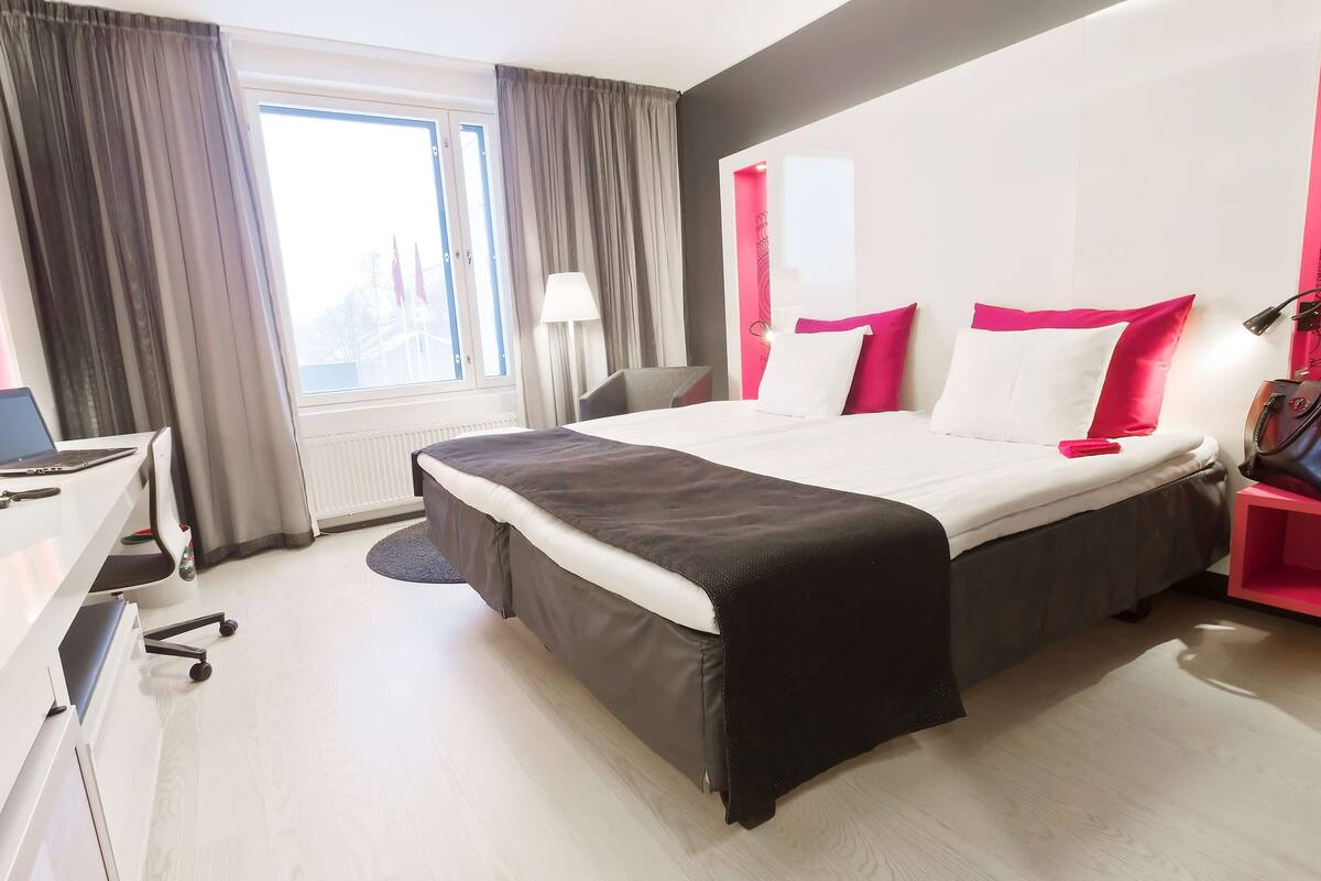 A modern hotel room with a large bed, a desk with a laptop, and a chair.
