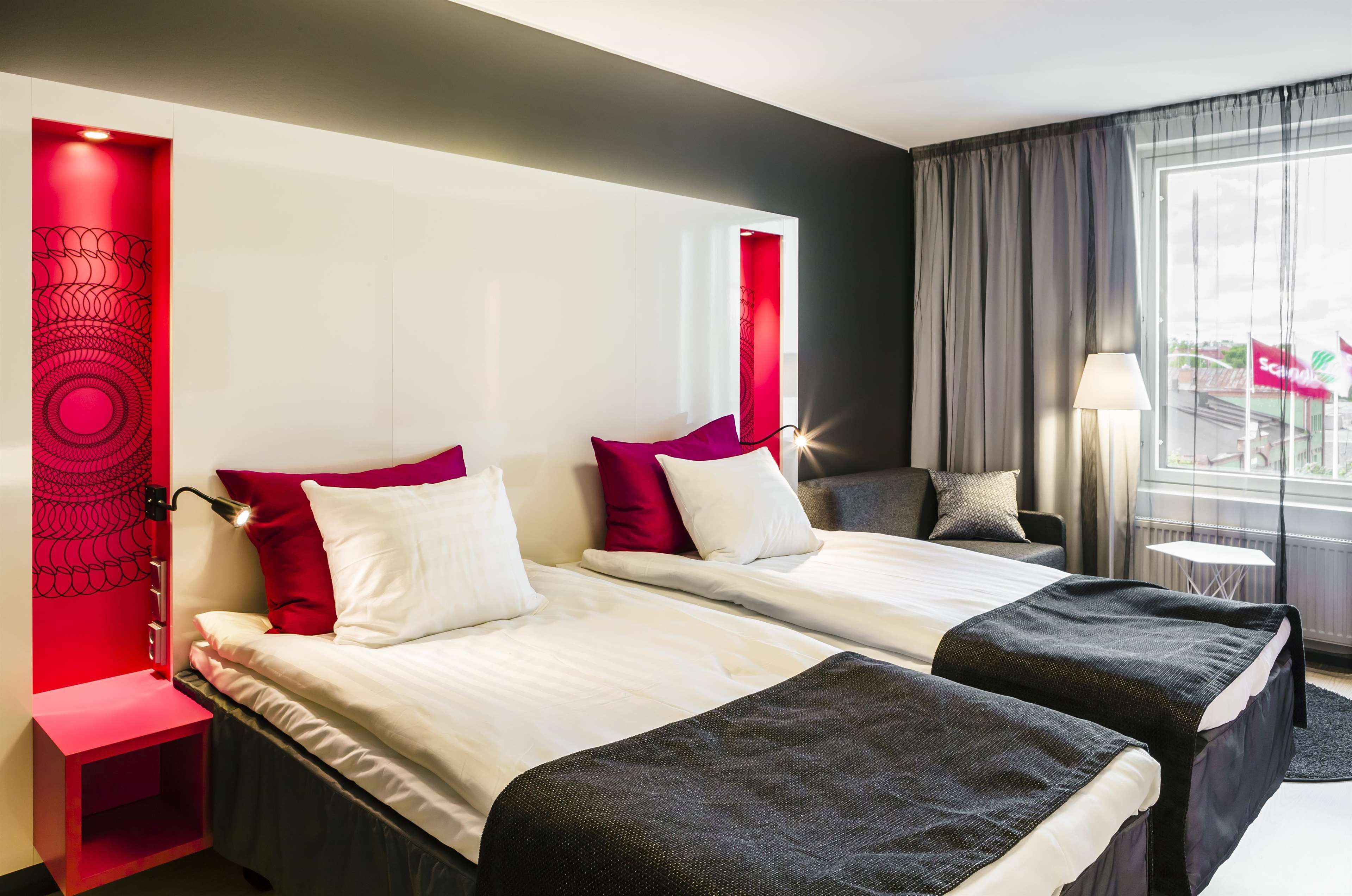 A hotel room with two beds, a red accent wall, a small table, and a window with curtains.