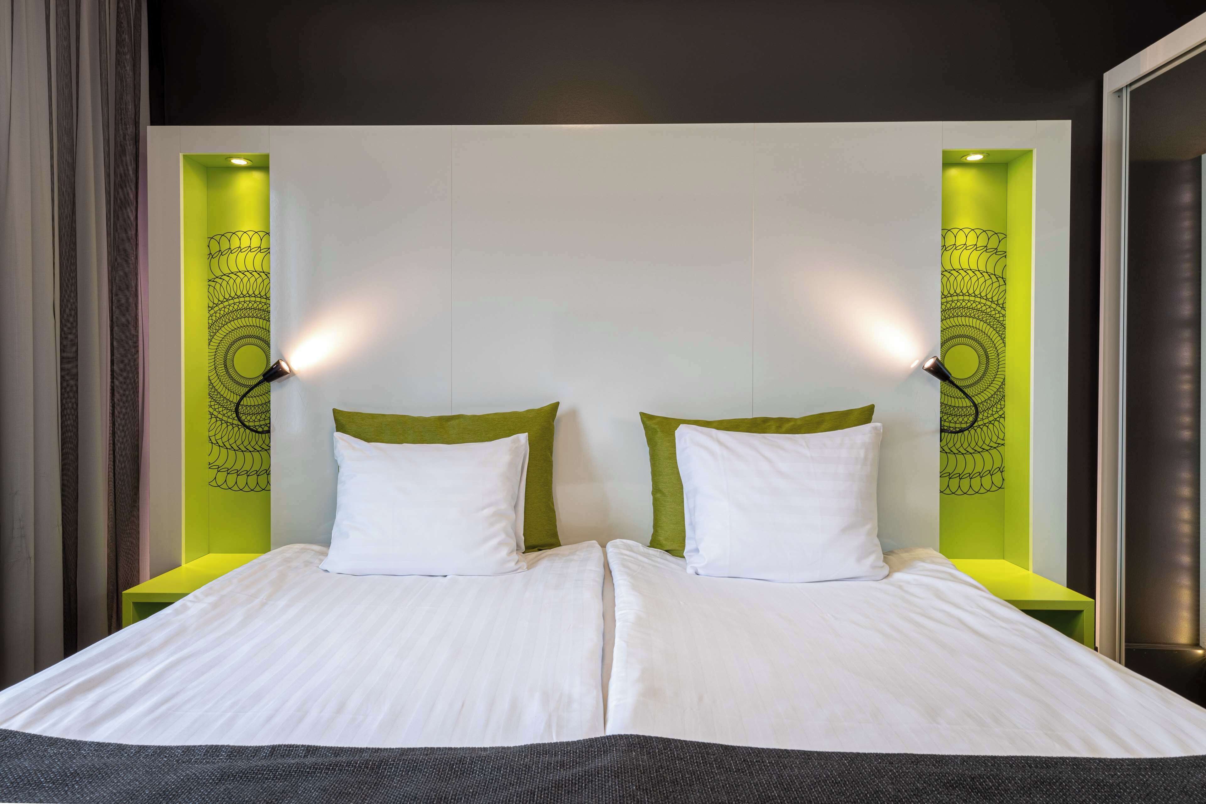 A double bed with white linens and green headboards, two bedside lamps, and a wall with a circular design.