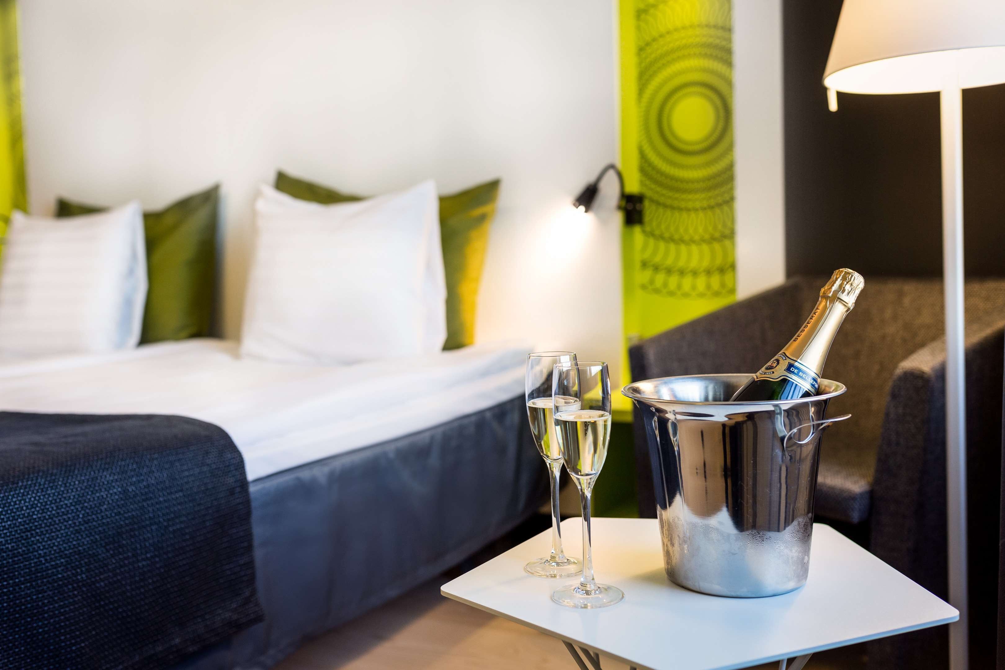 A hotel room with a bed, a small table with two glasses of champagne and a bottle in a bucket, and a lamp.