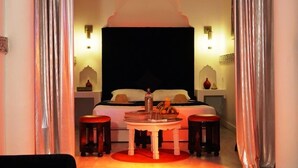 Una habitación con una cama, una mesa con una cesta de frutas, dos taburetes y apliques decorativos en la pared.