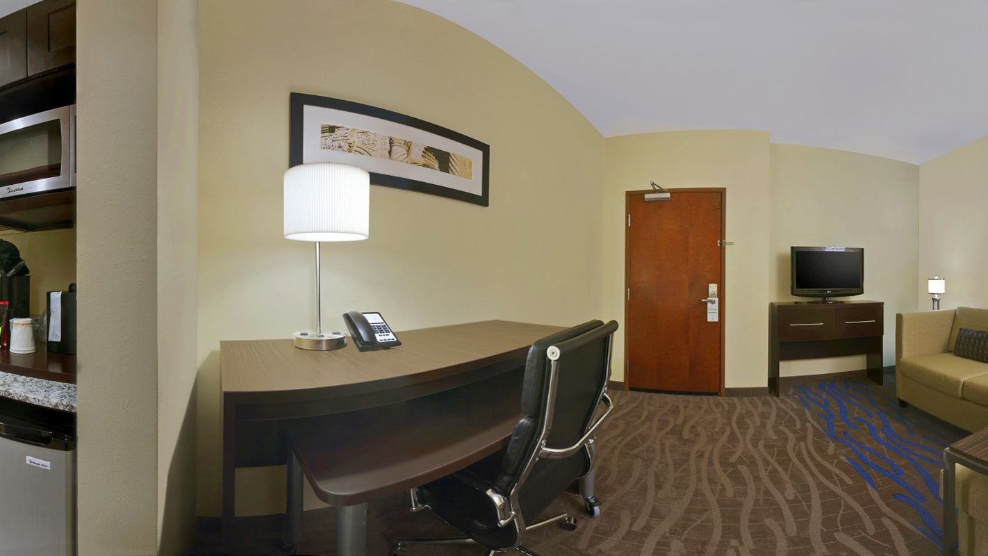 A hotel room with a desk, chair, TV, sofa, and a door.