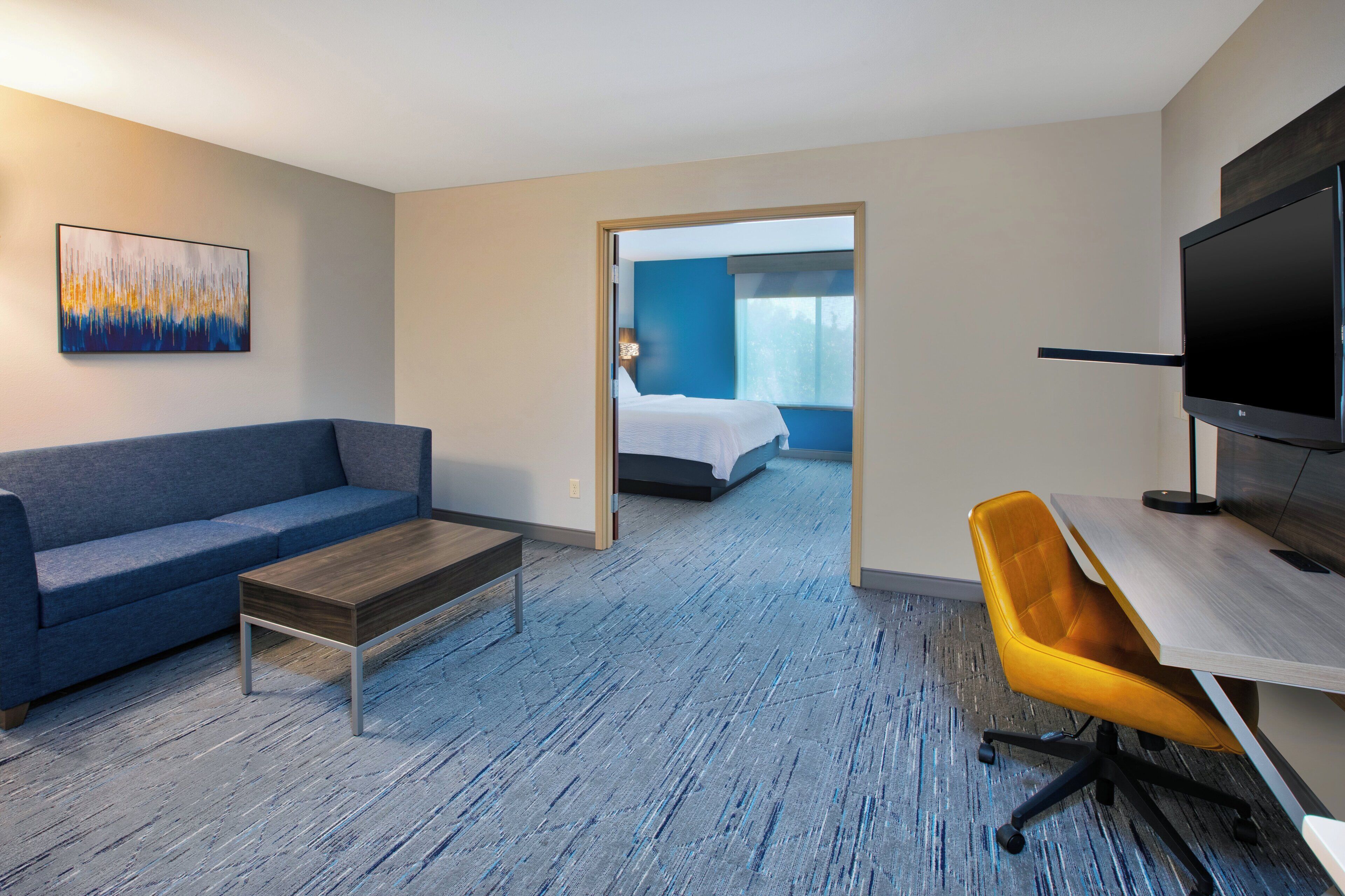 A hotel room with a blue sofa, a wooden coffee table, a yellow office chair, a flat-screen TV, and a bed with a blue headboard.