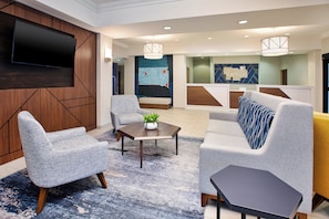A modern hotel lobby with a large flat-screen TV mounted on the wall, a seating area with sofas and armchairs, a coffee table with a plant, and a map mural on the wall. - Holiday Inn Express and Suites Sioux Falls SW by IHG (Sioux Falls)