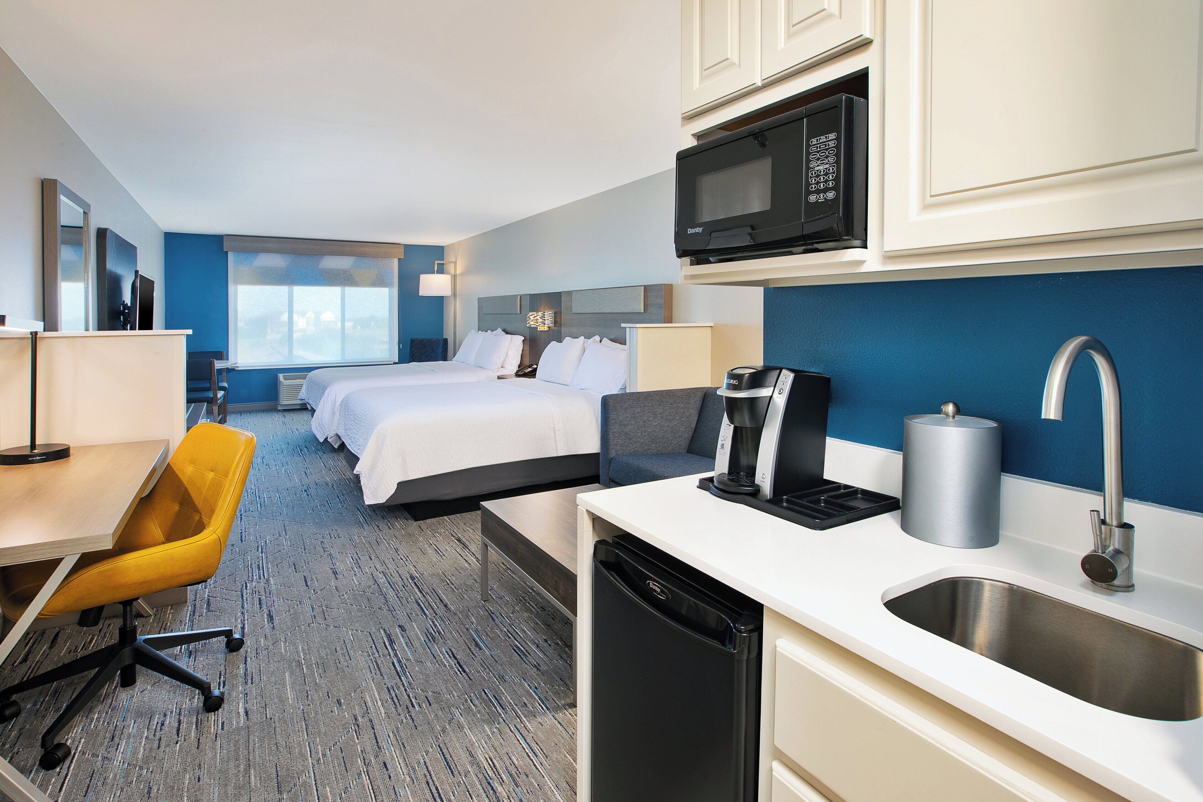 A hotel room with a bed, a desk, a chair, a microwave, a sink, and a coffee maker.